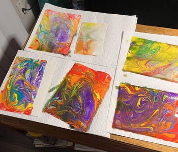 Handmade marbled paper sheets with vibrant, swirling patterns are laid out on paper towels to dry on a wooden table