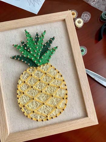 Pineapple-themed string art in a wooden frame displayed on a table, with tools and threads nearby