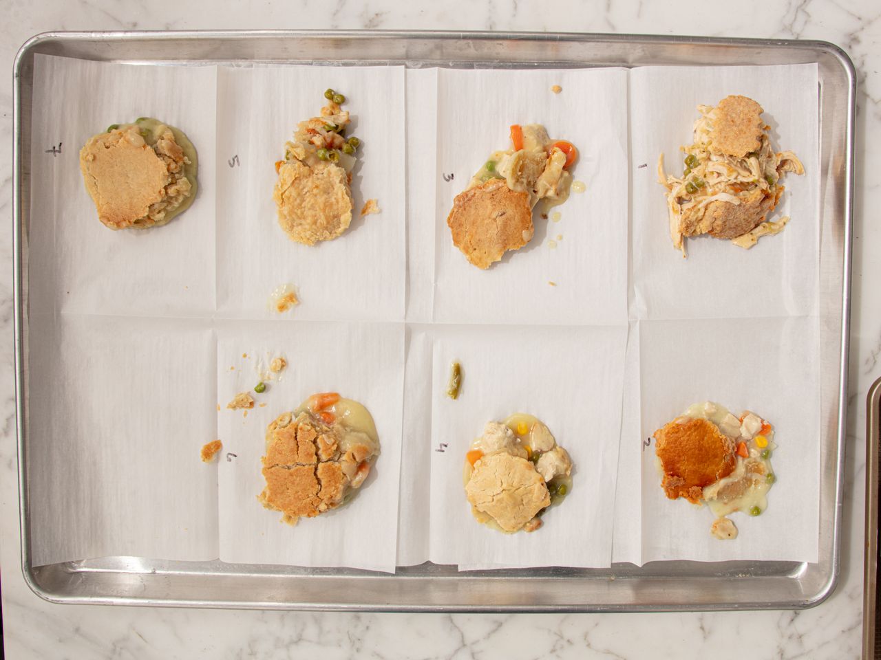 Eight samples of chicken pot pies on a baking tray arranged on separate parchment sheets with visible fillings and toppings