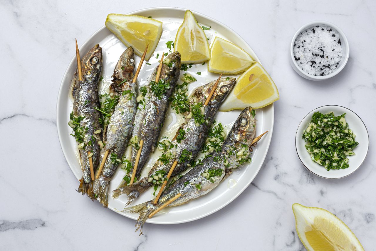 A plate of grilled whole sardines garnished with chopped herbs and lemon wedges accompanied by small dishes of herbs and salt on the side