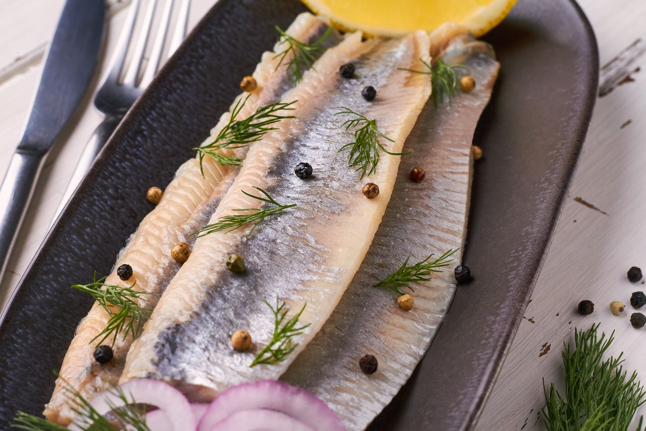 Three fillets of fish served on a black platter garnished with dill and peppercorns with a lemon wedge and sliced onion on the side
