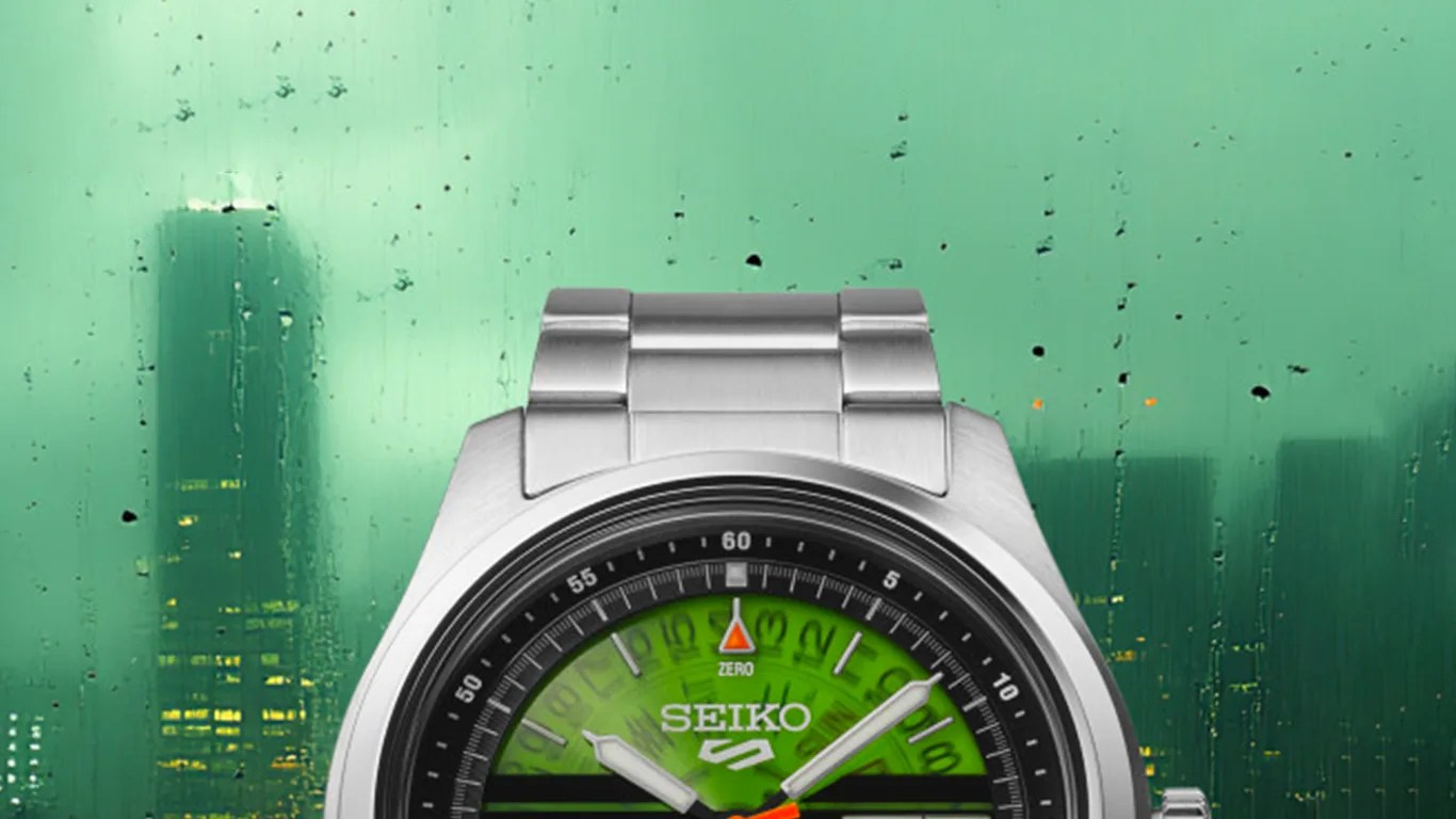 Silver Seiko wristwatch with a green dial and metal link bracelet against a rainy cityscape background.
