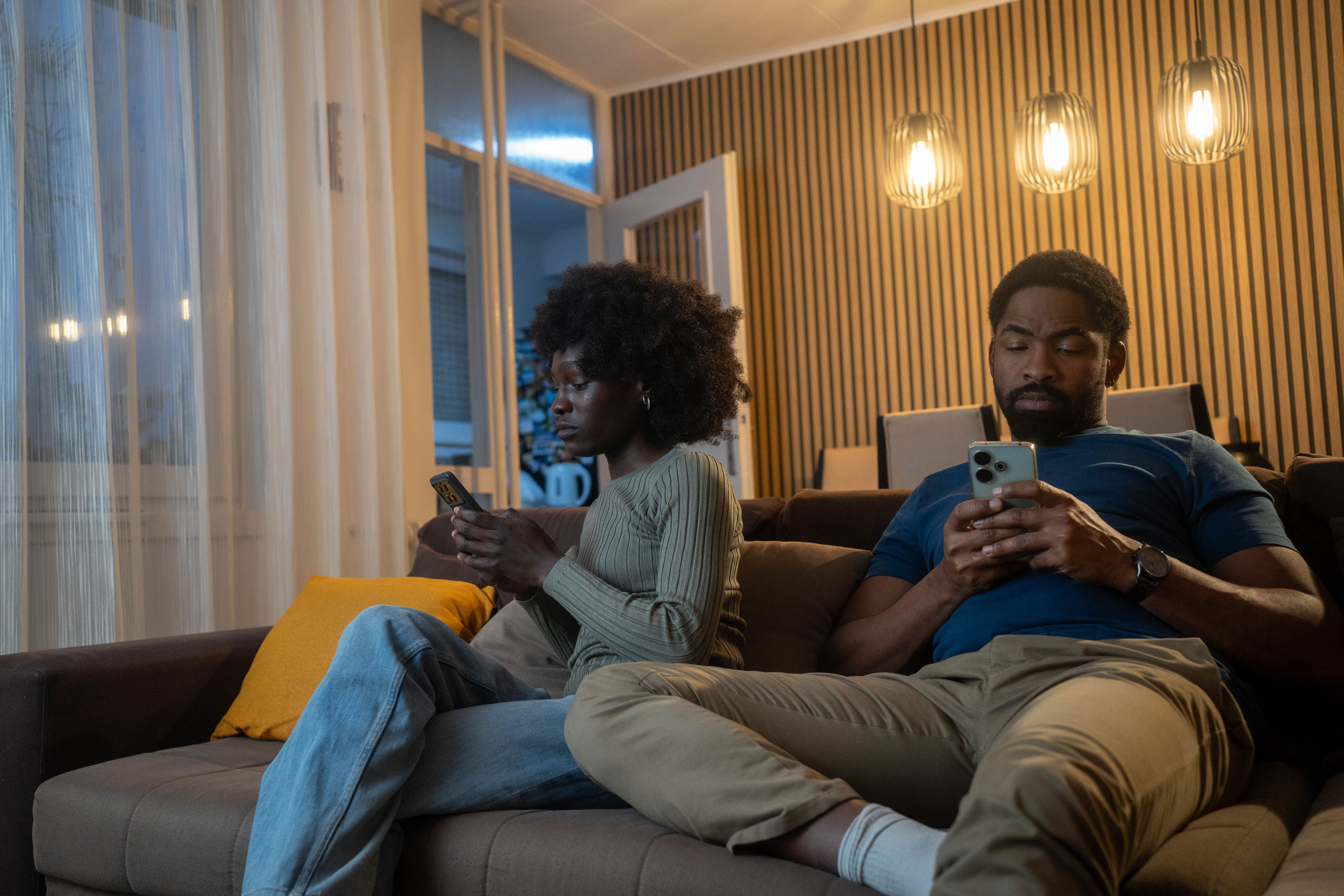 Two people sit on a couch, focused on their phones, in a living room with modern pendant lights and wood paneling