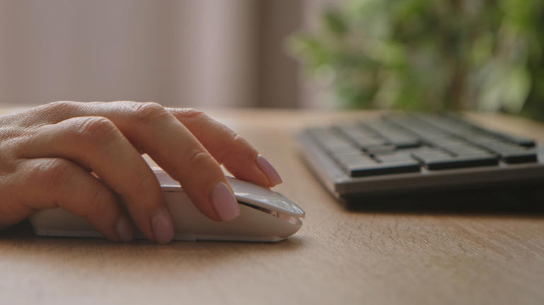 A woman's hand operates a mouse on a desk next to a keyboard.