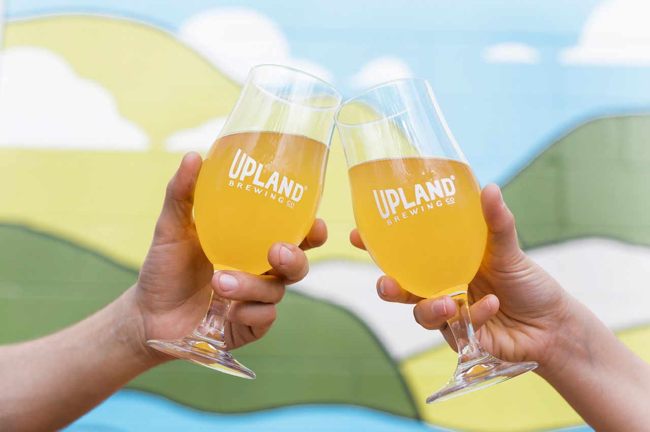 Two hands holding glasses of beer with Upland Brewing logo raised in a toast against a background of painted hills and sky