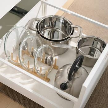 Organized kitchen drawer with stainless steel pots, a kettle, and glass lids on a wooden rack