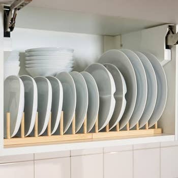 Neatly organized white plates and bowls are stacked vertically on a wooden dish rack inside a kitchen cabinet