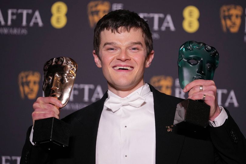An individual in formal attire, wearing a tuxedo, proudly displays multiple awards, with a backdrop featuring the logo and branding of a prestigious event.