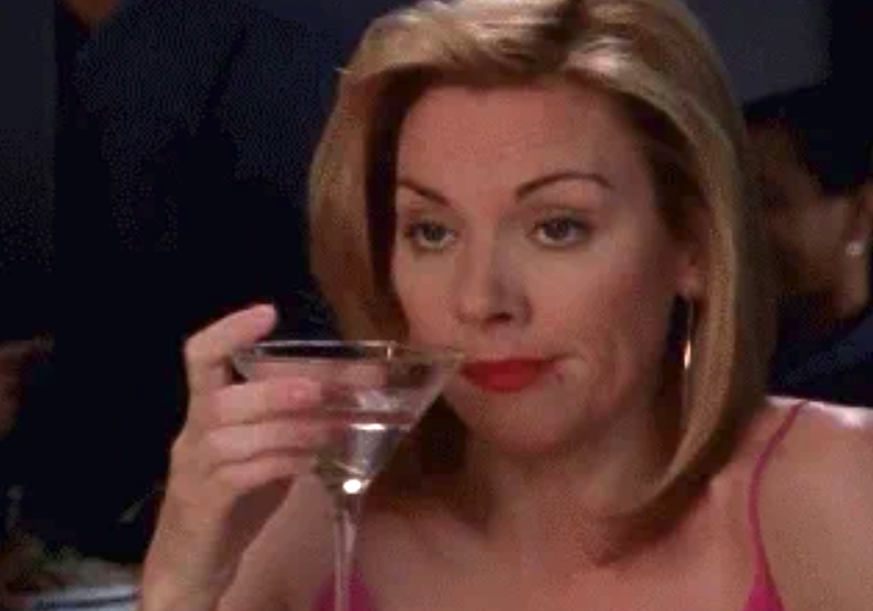 A person sips from a martini glass with a contemplative expression, sitting at a table in a casual setting
