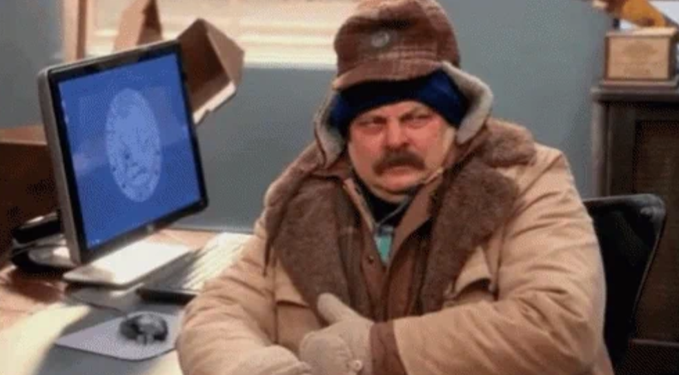Person bundled in winter gear sitting at office desk, looking displeased. Computer monitor in background