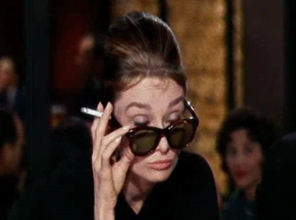 A person wearing sunglasses indoors, holding a cigarette, with an expression suggesting curiosity or surprise