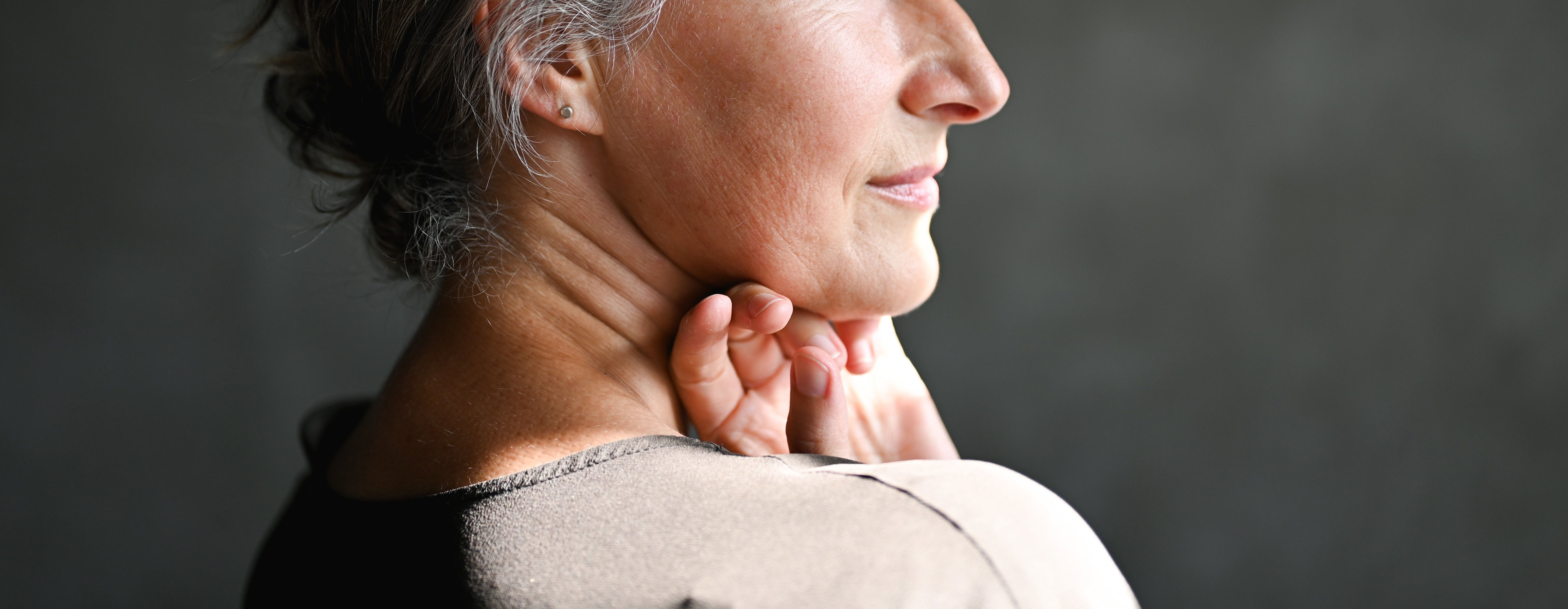 Elderly woman with short, gray hair gently touching her neck, gazing thoughtfully into the distance