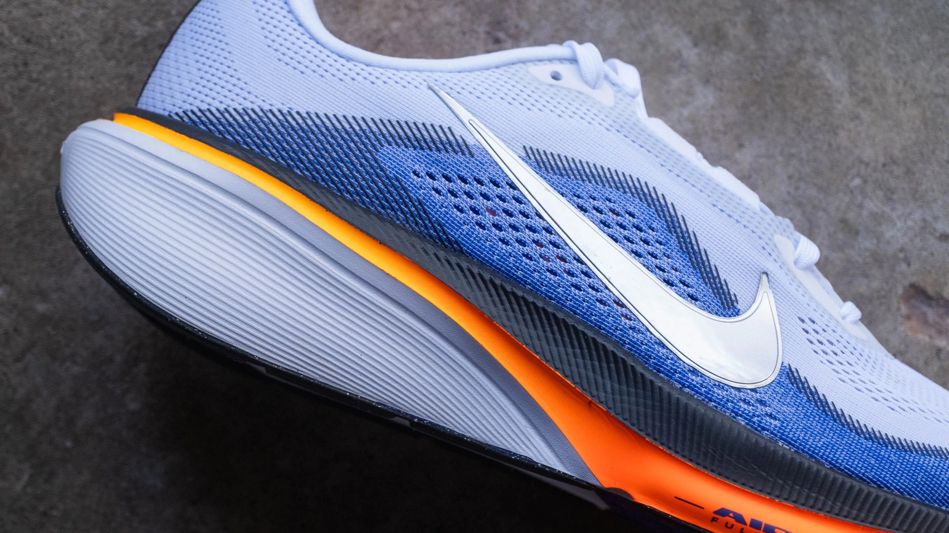White and blue Nike running shoe with orange midsole and large white swoosh logo on a gray surface.