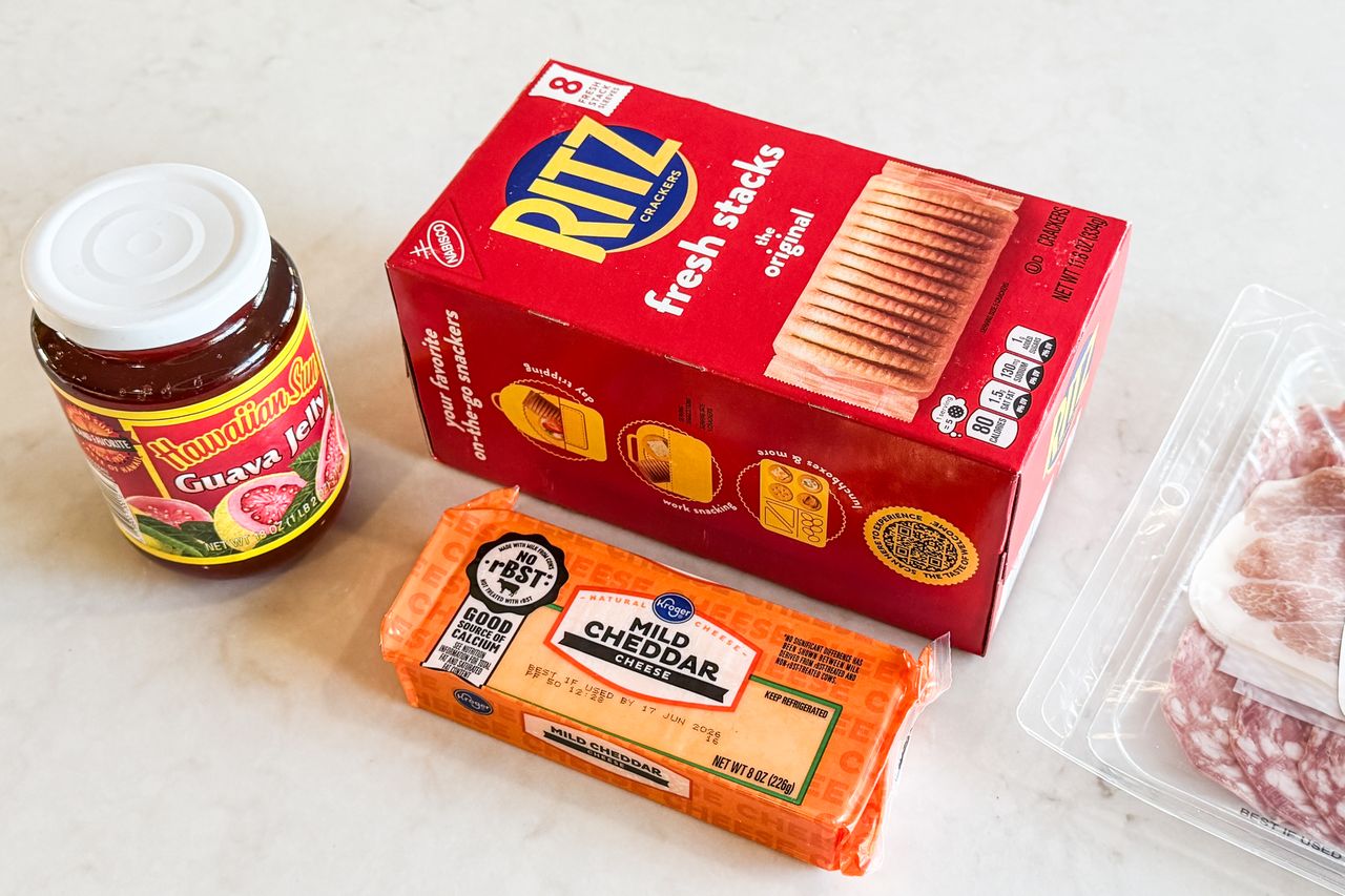 A jar of guava jelly a box of Ritz crackers a block of mild cheddar cheese and a package of deli meat placed on a counter