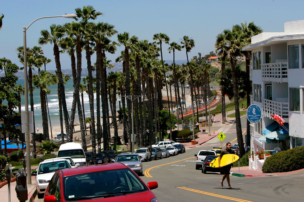 A beachside street with palm trees parked cars and a person carrying a surfboard