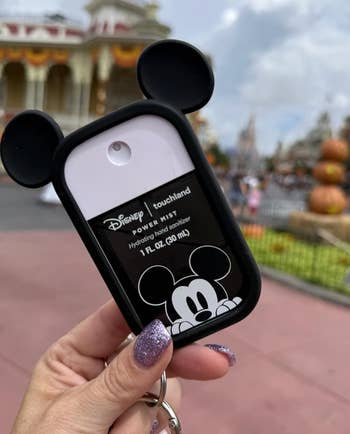 Hand holding a Disney-themed hand sanitizer with Mickey Mouse ears, set in an amusement park background with pumpkins