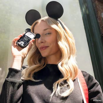 model with mouse ears holds Mickey-themed skincare product near face, smiling. Wearing a black top, featured in a shopping article