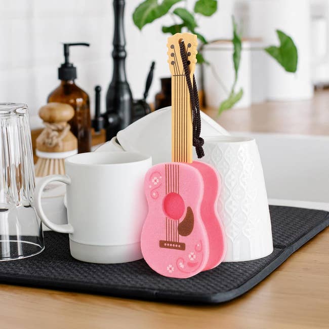 Pink guitar-shaped kitchen sponge on a dish rack with cups and plates