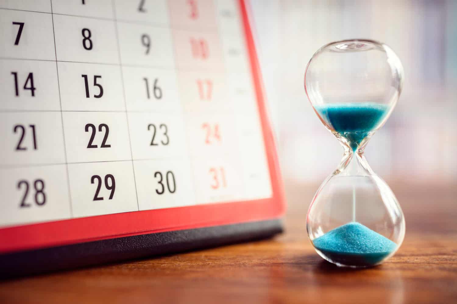 A close-up shot shows a white calendar page with black numbers, partially obscured, next to a clear glass hourglass containing bright blue sand. The majority of the blue sand is still in the upper bulb of the hourglass, with a small pile already accumulated in the lower bulb, suggesting time passing. The objects rest on a warm brown wooden surface.
