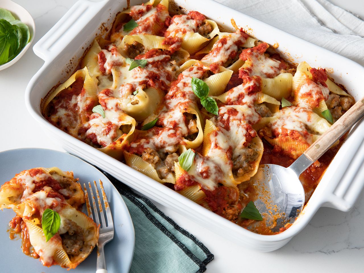 close up view of Italian Sausage Stuffed Shells garnished with fresh basil in a white baking dish with a serving spoon and Italian Sausage Stuffed Shells on a blue plate