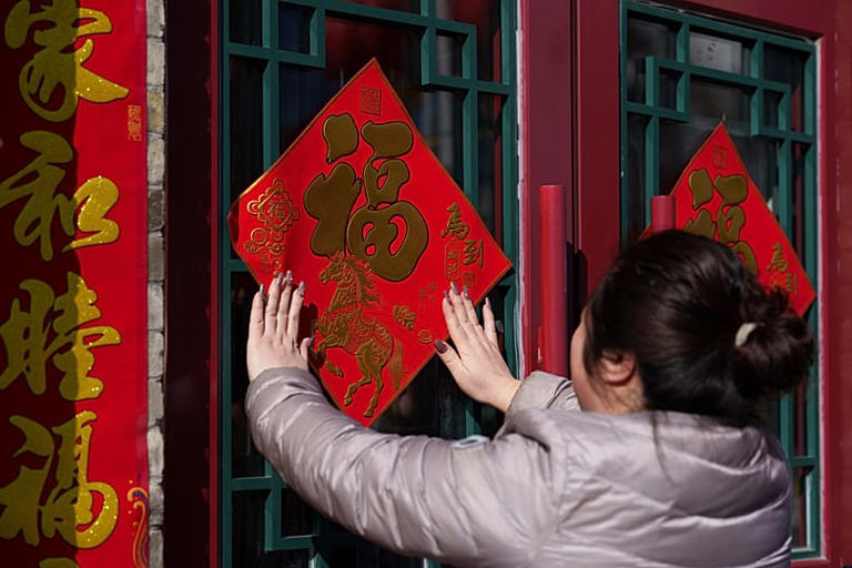 Una mujer pega una decoración de caracteres 'Fu' en una puerta antes del Año Nuevo Lunar en Pekín, China, el lunes 16 de febrero de 2026. (AP Photo/Vincent Thian) Vincent Thian/Copyright 2026 The AP. All rights reserved