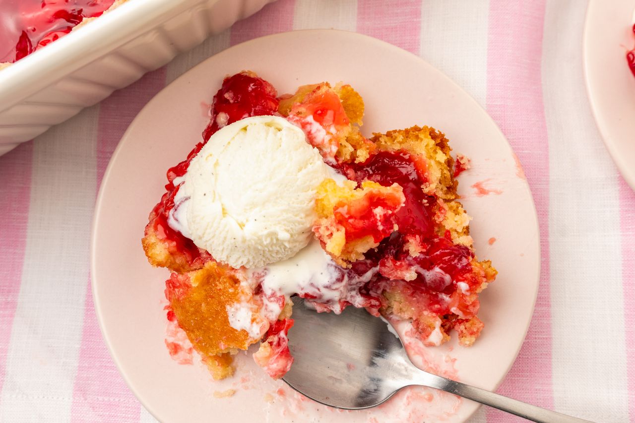 A slice of cherry dump cake topped with a scoop of ice cream on a plate served with a spoon