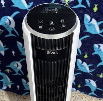 Levoit tower fan on a patterned carpet with a shark-printed backdrop, featuring touch controls on top