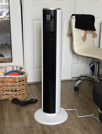 A vertical tower fan in a cluttered room with a woven basket and swivel chair nearby