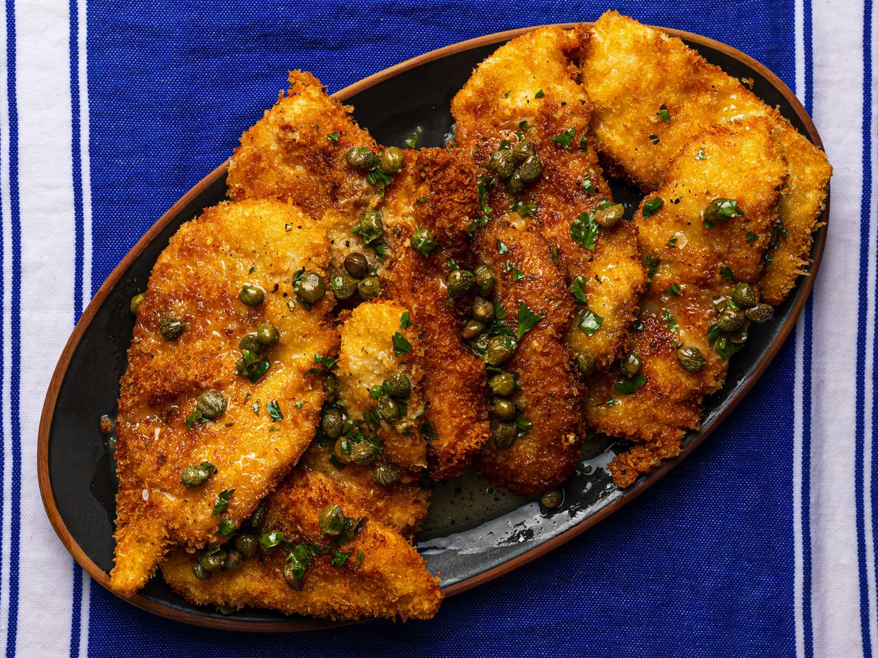 An oval platter holding fried chicken cutlets topped with lemon-butter pan sauce containing capers. The platter is on a blue and white dish towel.