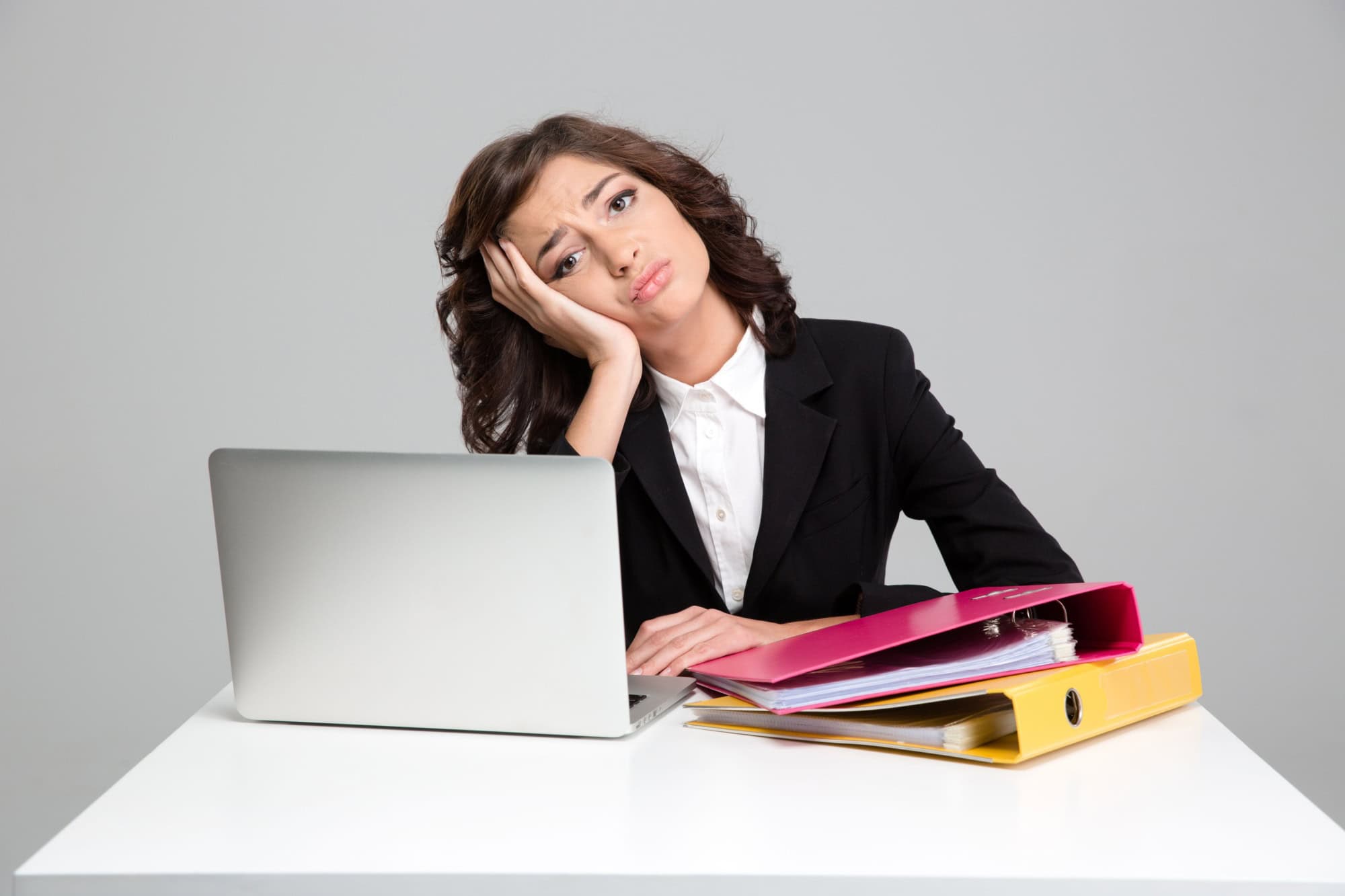 Bored sad depressed curly pretty young woman using laptop and working with documents in colorful binders