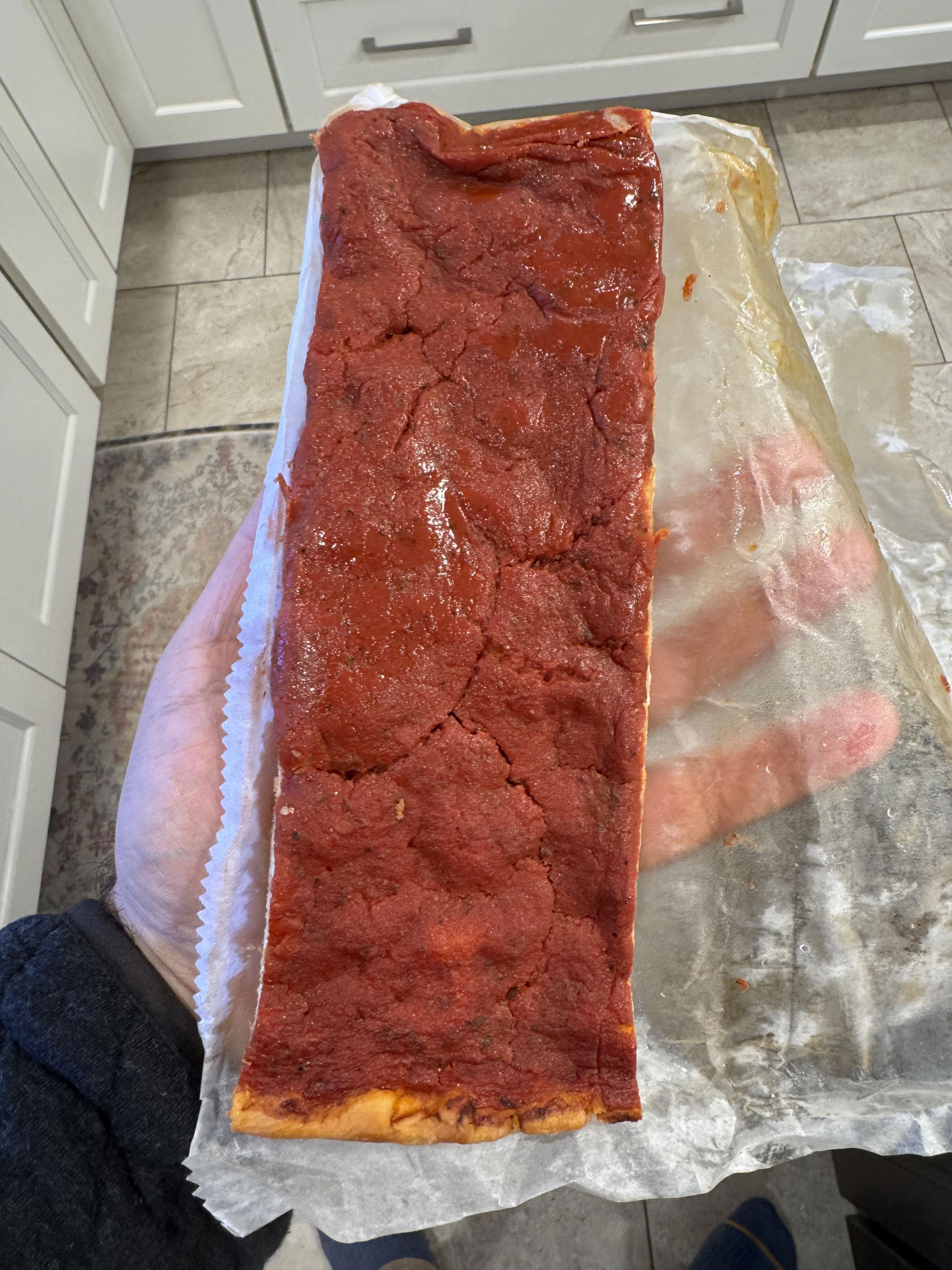 A hand holds a rectangular pizza slice with tomato sauce on a parchment sheet, in a kitchen setting