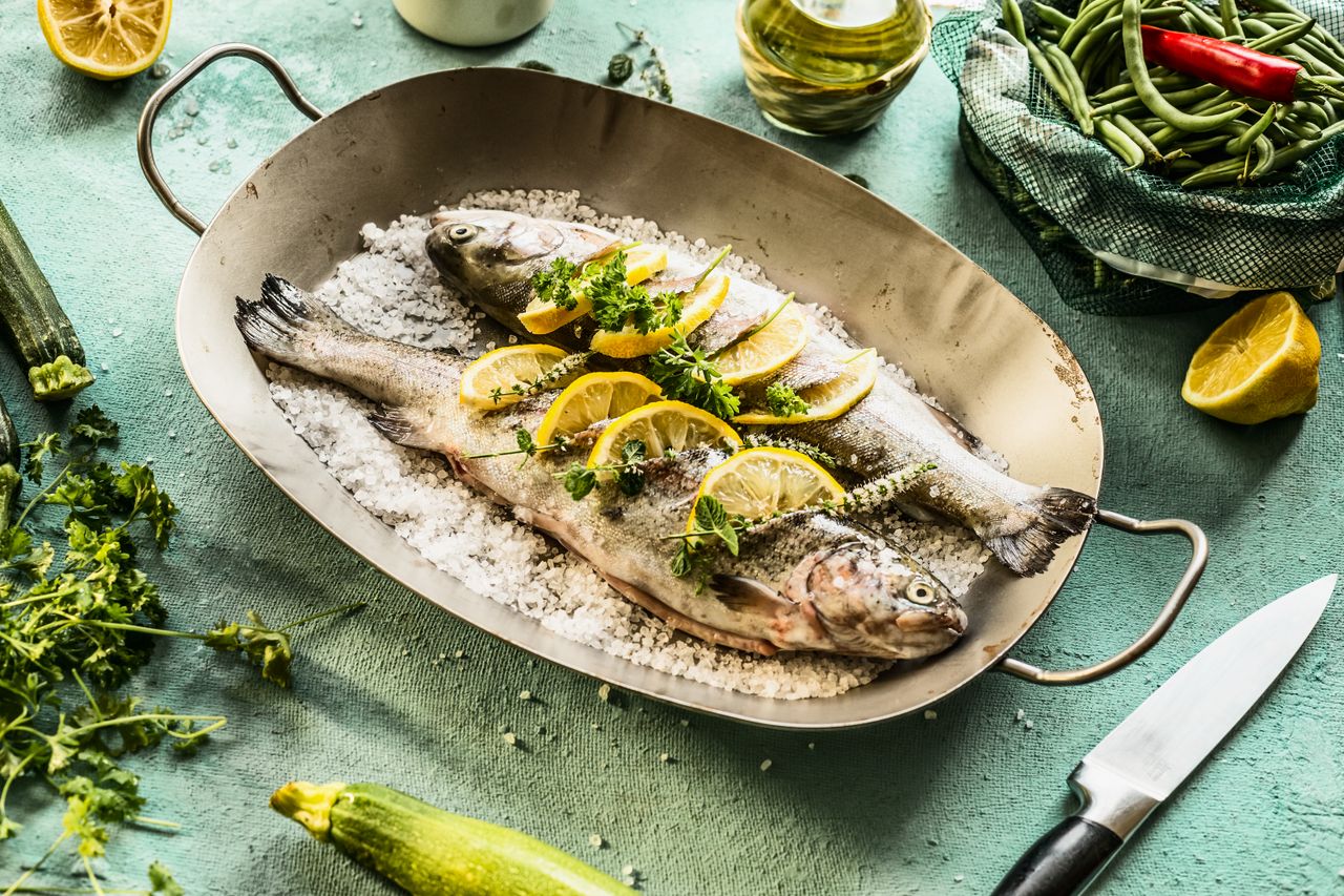 Two trout stuffed with herbs and lemon slices in fish pan on kitchen table with herbs, spices and kitchen utensil. Real fish cooking preparation scene. Step by step