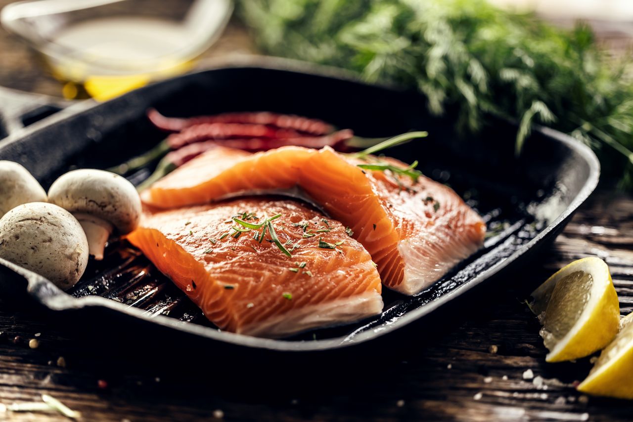 Two raw salmon fillets ready to be grilled in a black cast iron skillet. Various ingredients, such as fresh dill and rosemary, lemon slices, chili peppers, mushrooms, and olive oil, are arranged around the skillet on a dark wooden surface, suggesting the preparation of the dish.