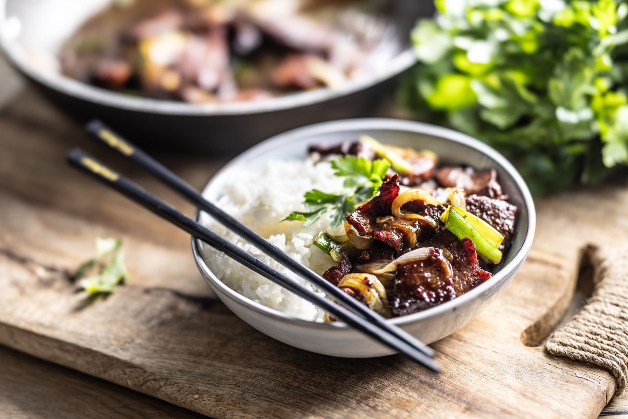 A close-up of a bowl of Asian food placed on a wooden board. The bowl contains white rice and stir-fried beef with vegetables such as green onions and caramelized onions, garnished with fresh parsley. A pair of black chopsticks rests on the side of the bowl.