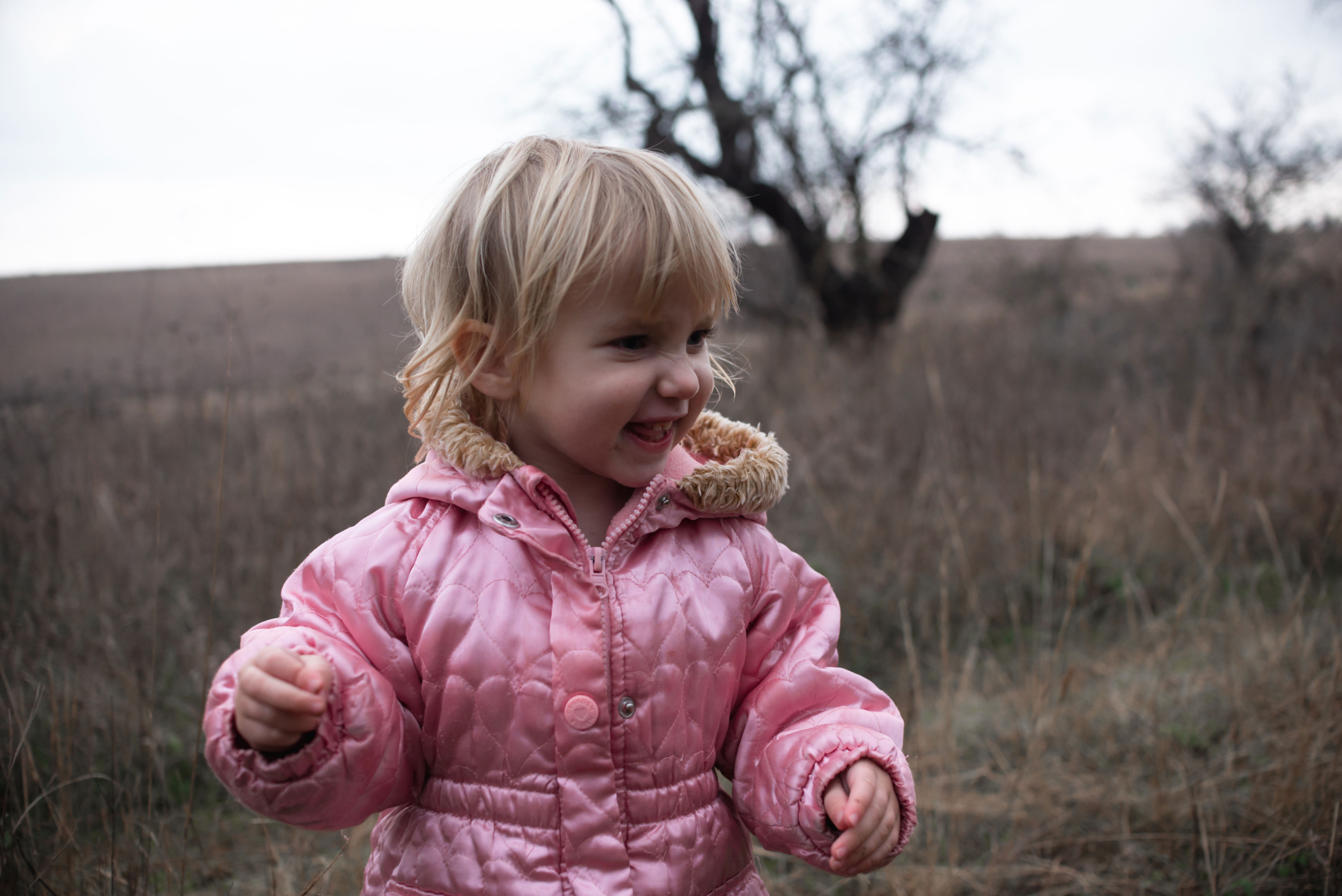 Young child smiling in a field, wearing a puffy jacket. The landscape is grassy with bare trees in the background