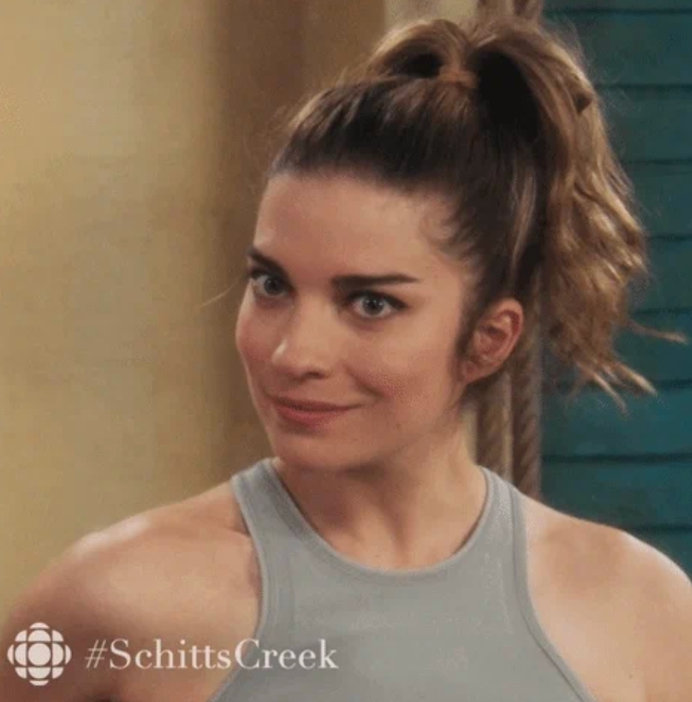 Woman with high ponytail giving a sly smile, wearing a sleeveless top. "#SchittsCreek" is visible in the corner