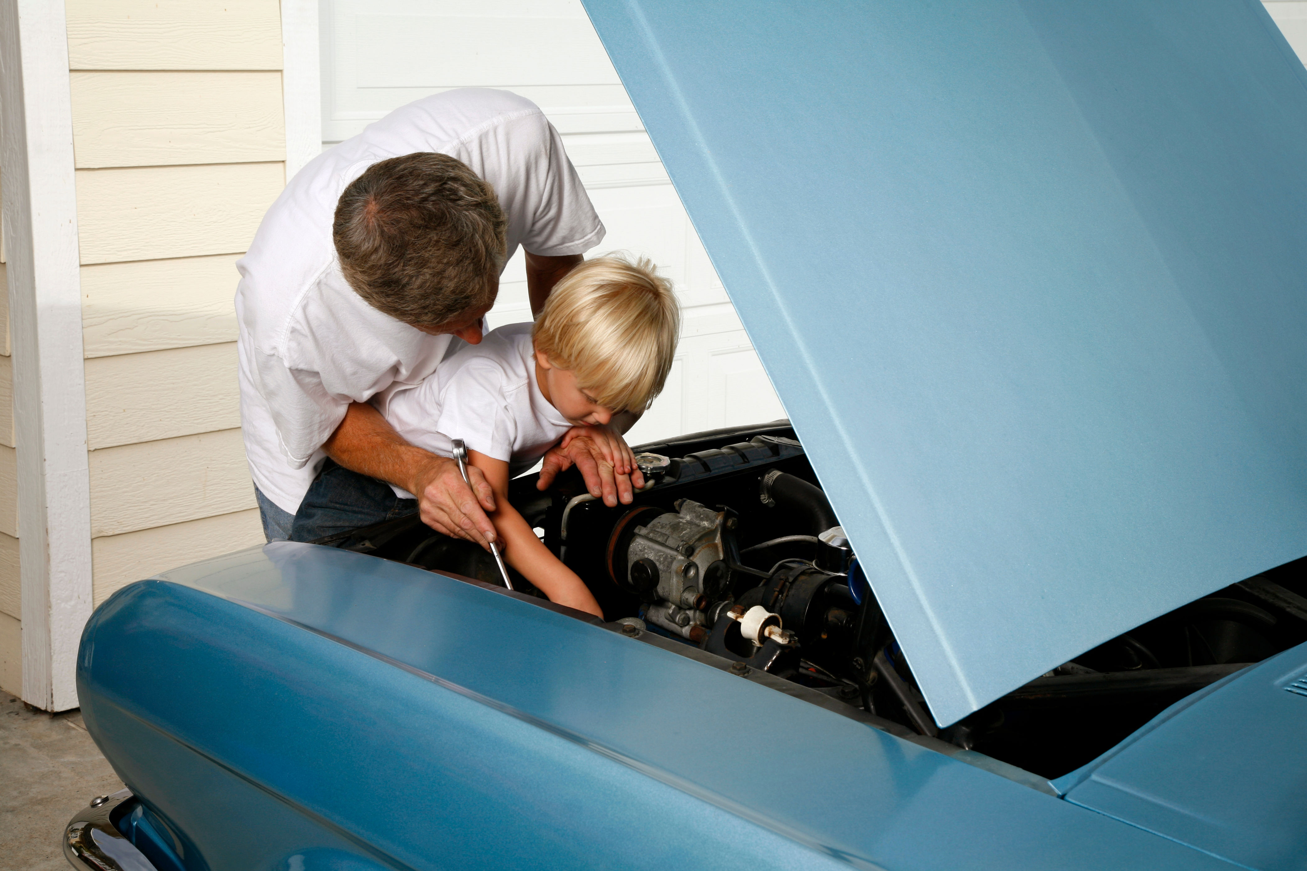 Man helps child inspect car engine, both focused under open hood in a garage setting