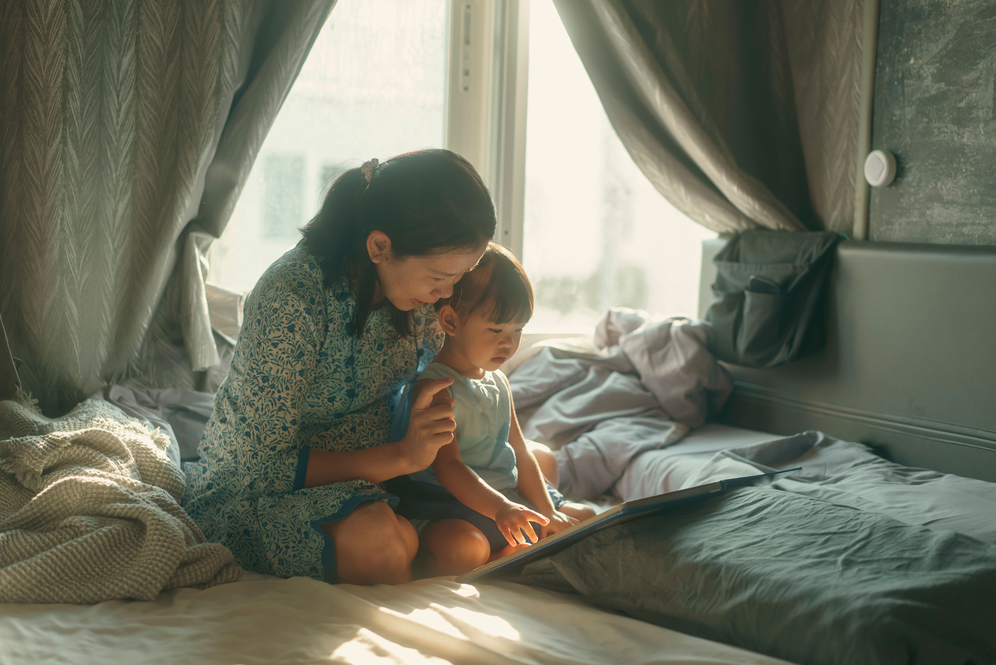 A mother and child sit on a bed, reading a book together by the window, wrapped in a cozy atmosphere