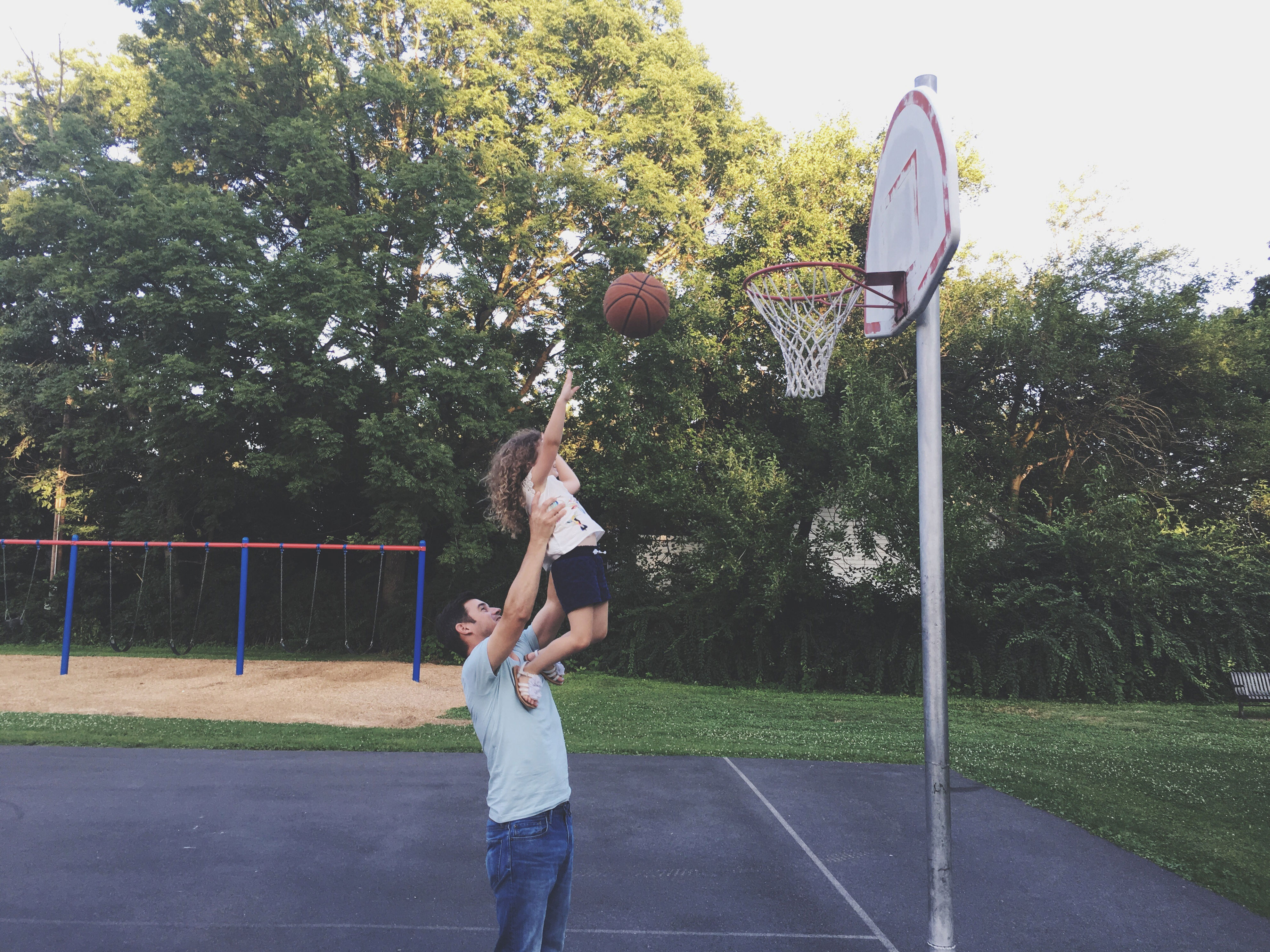 A man lifts a child at a basketball hoop, helping them dunk a basketball on an outdoor court. Trees and a playground are in the background