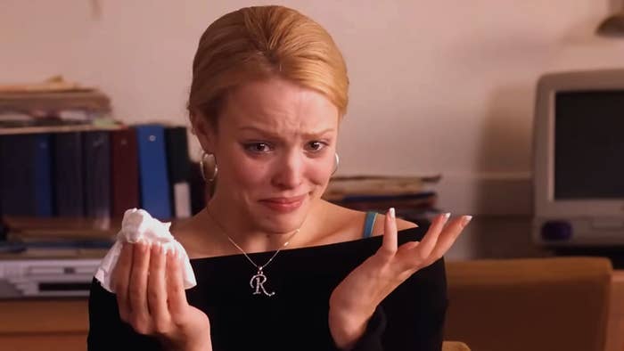 Person with blonde hair crying and holding tissues, wearing a black top with a necklace. Office setting in the background