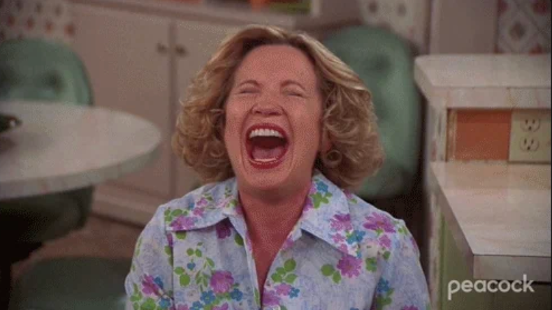 A woman laughing heartily in a kitchen setting, wearing a floral blouse, with an expression of joy