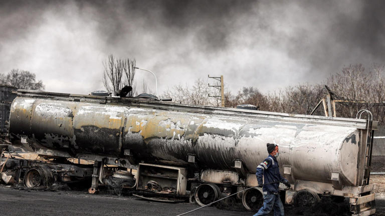 An Iranian civil defence member walks with a hose next to a destroyed fuel tanker vehicle near an ongoing fire following an overnight airstrike on the Shahran oil refinery in northwestern Tehran on March 8, 2026.