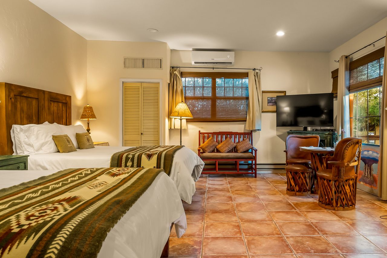 A cozy guest room with two queen beds a small sofa a table with chairs a TV and a window with natural light shining through