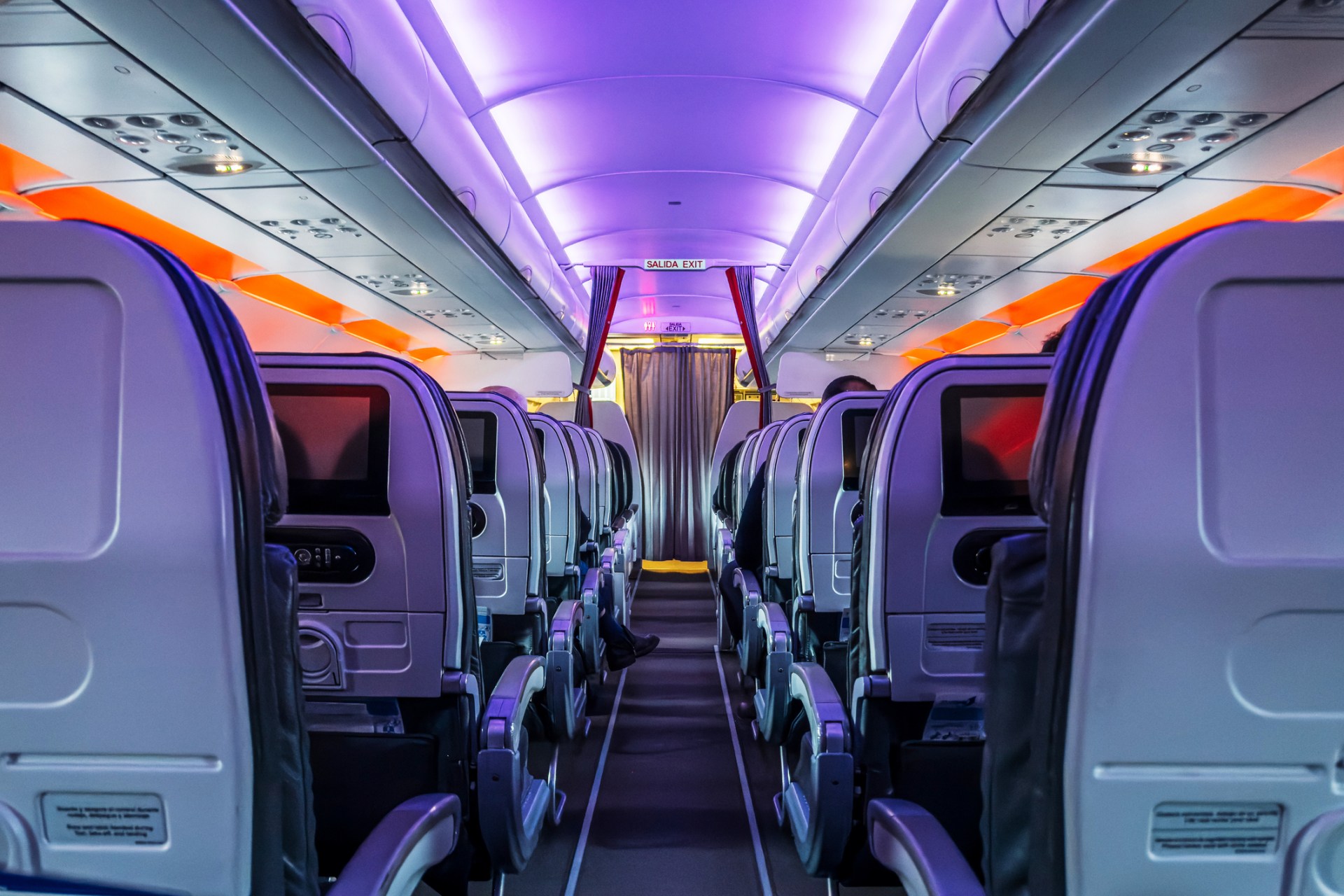 A plane cabin with the aisle centred and seats on either side, lit-up with purple and orange lights during an evening flight.