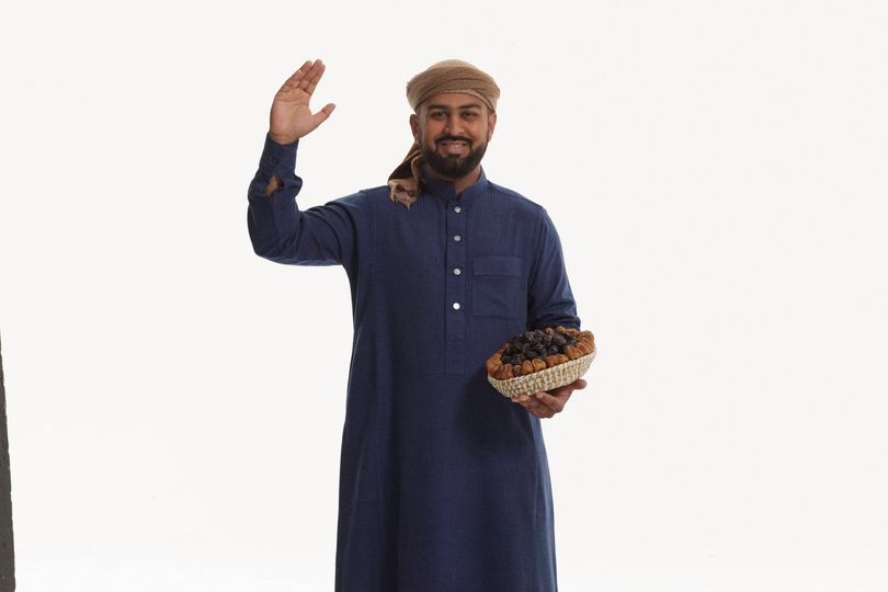 An individual dressed in a dark blue garment, resembling a traditional robe, stands with one hand raised and the other holding a bowl of dates, against a plain, light-colored background.