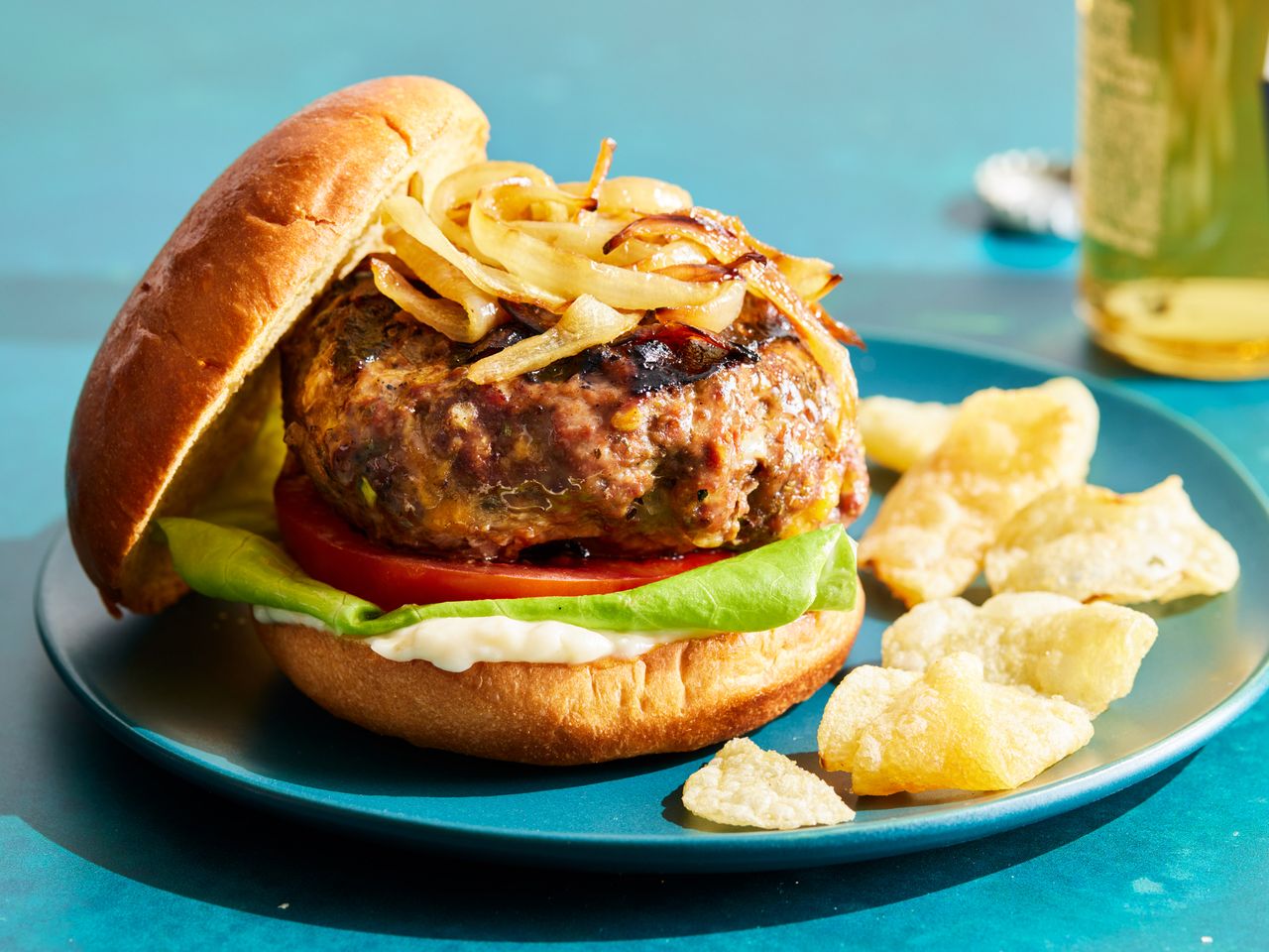 A hamburger served on a bun with fried onions, lettuce, and tomato, served with potato chips