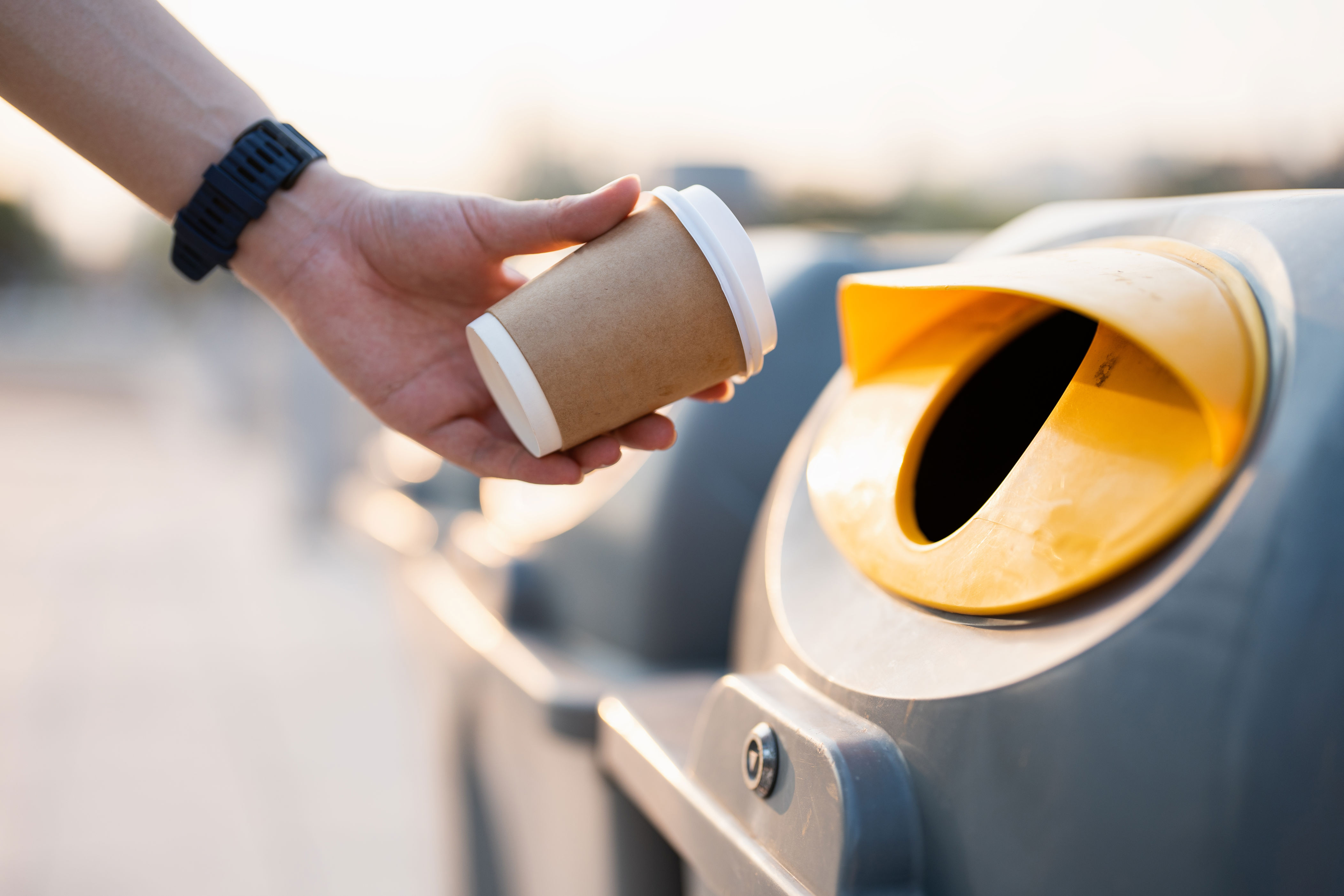 A hand holding a disposable cup near a recycling bin, symbolizing environmental awareness and sustainability in workplace practices