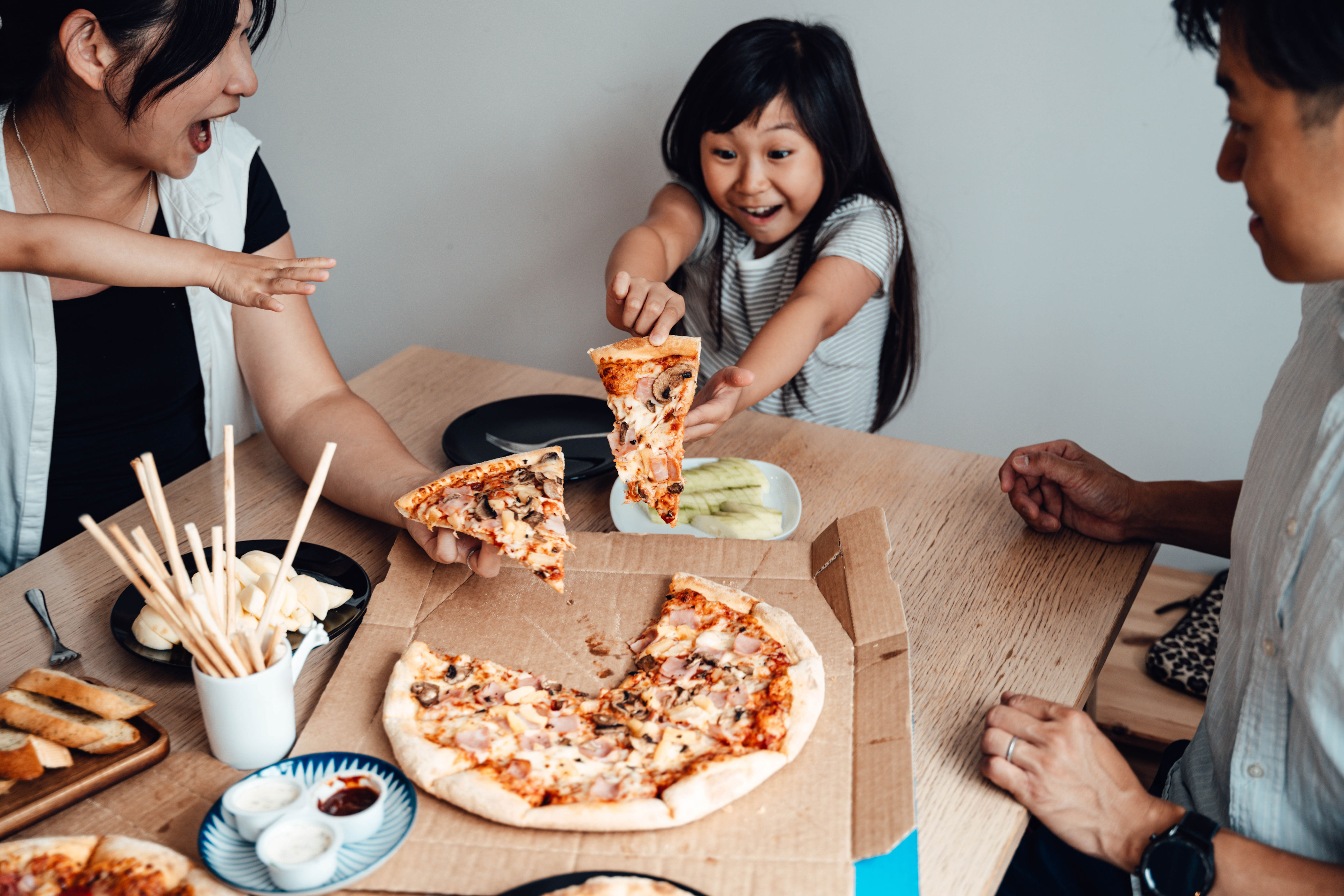 Family enjoying pizza around a table, with a young girl excitedly reaching for a slice