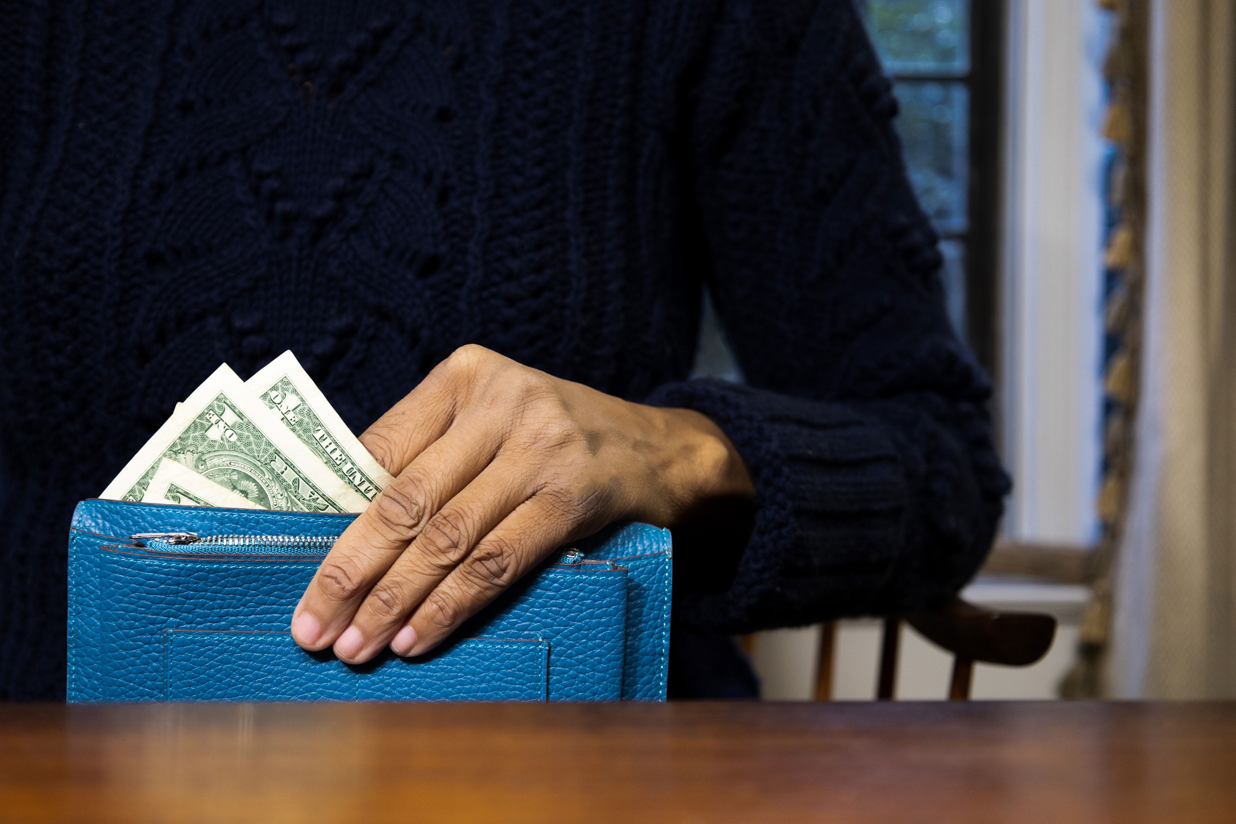 Person in knit sweater placing dollar bills into a blue wallet at a table, focusing on personal finance and money management