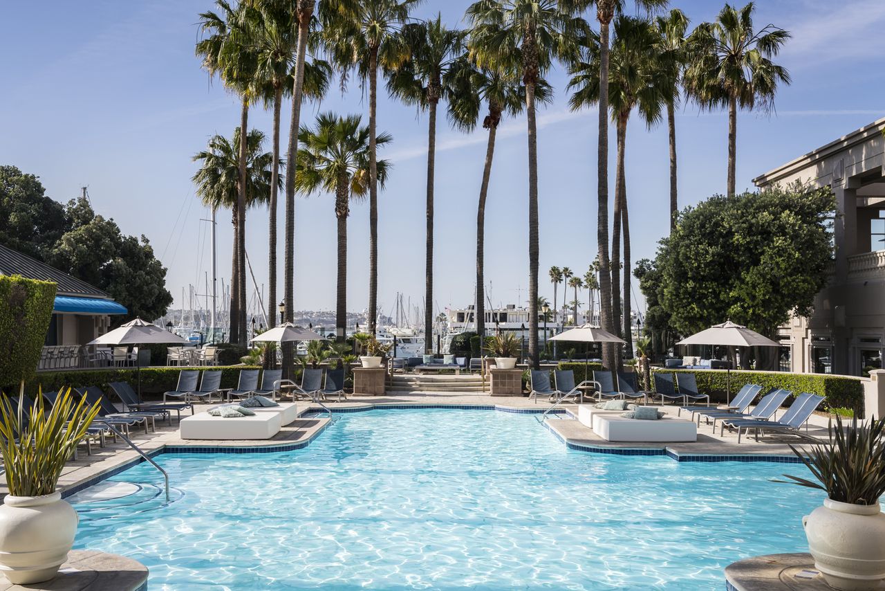 Outdoor swimming pool surrounded by lounge chairs and palm trees marina visible in the background